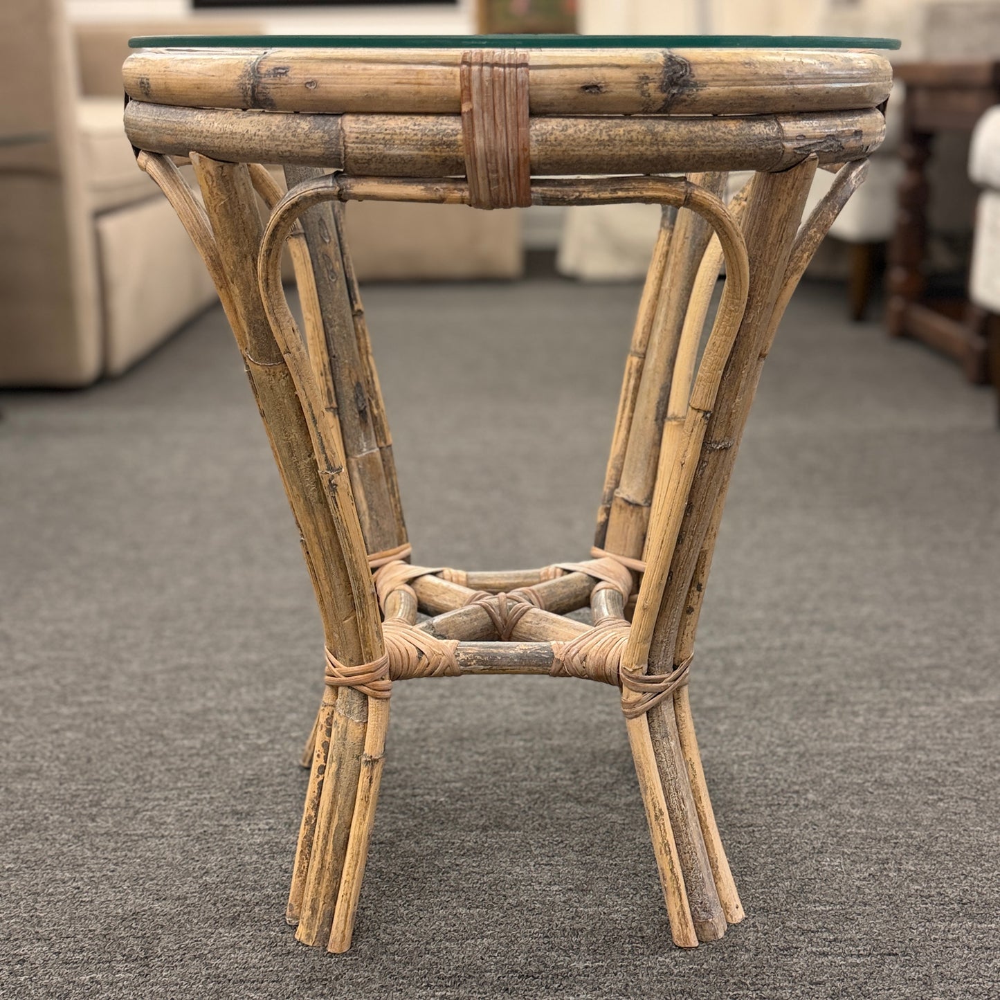 A small round end table with a bent organic-style bamboo frame, wrapped structural joints, an X-shaped cross-stretcher, and a clear glass tabletop.
