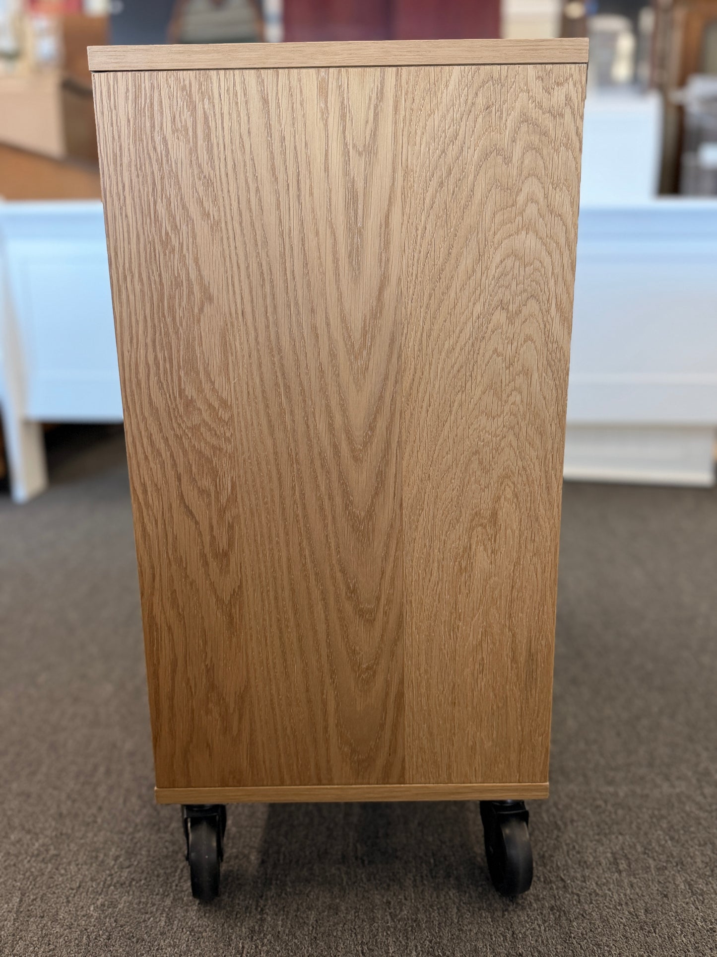 A modern mobile office storage cart with a light oak finish, featuring asymmetrical divided cubby compartments and four black rolling casters.