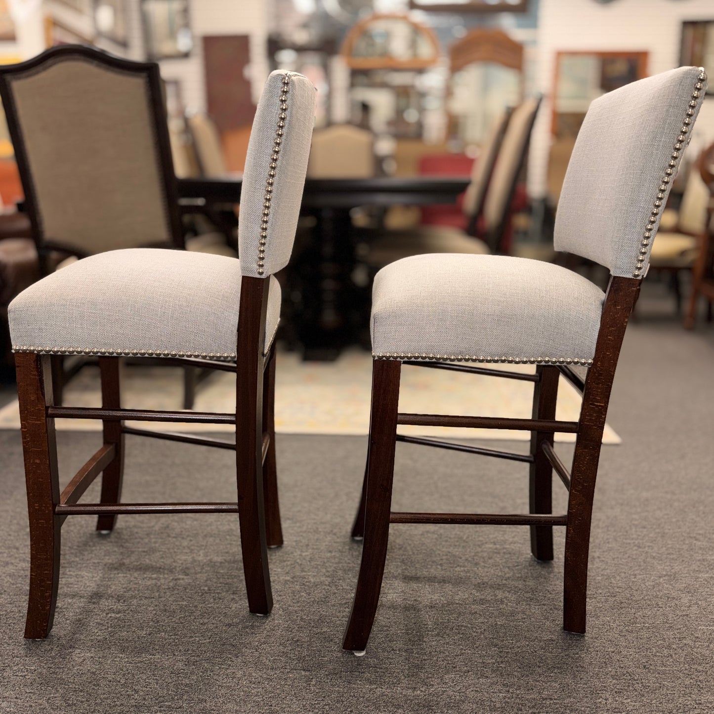 A pair of upholstered high-back counter stools featuring light grey textured fabric, classic metal nailhead trim, and dark-stained wooden legs with footrests.