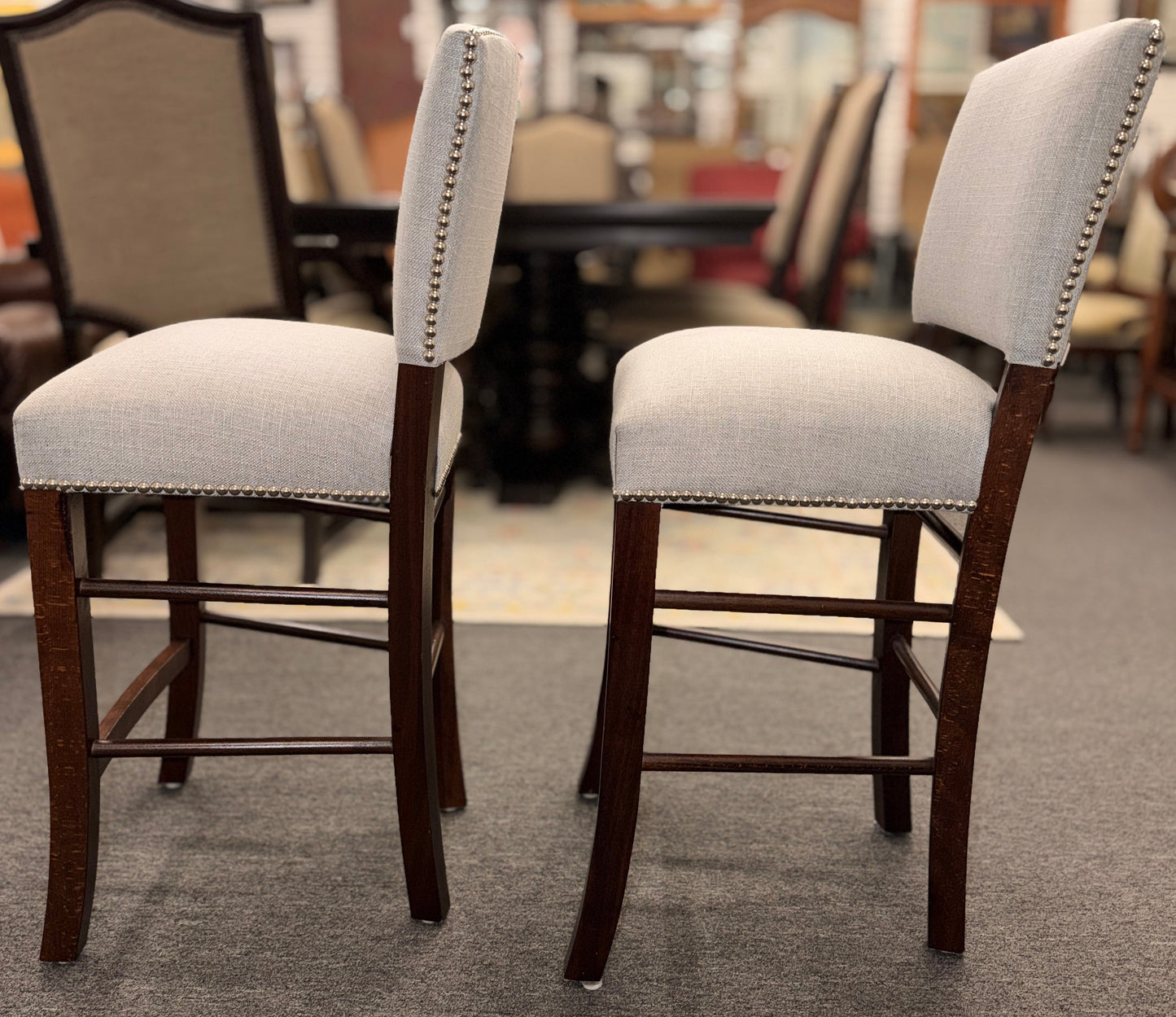 A pair of upholstered high-back counter stools featuring light grey textured fabric, classic metal nailhead trim, and dark-stained wooden legs with footrests.