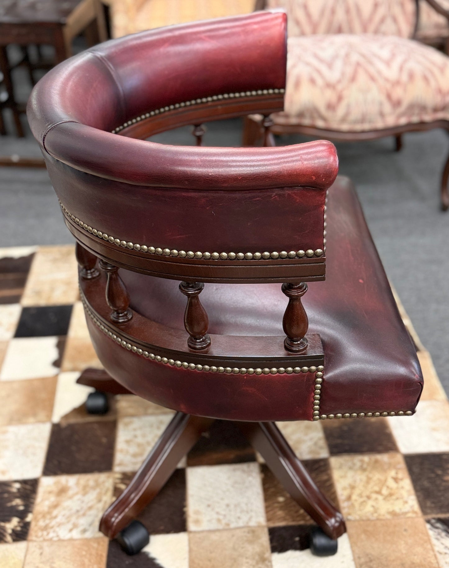A classic traditional rolling captain's desk chair featuring a curved back, wooden spindle supports, dark leather upholstery, and brass nailhead detailing.