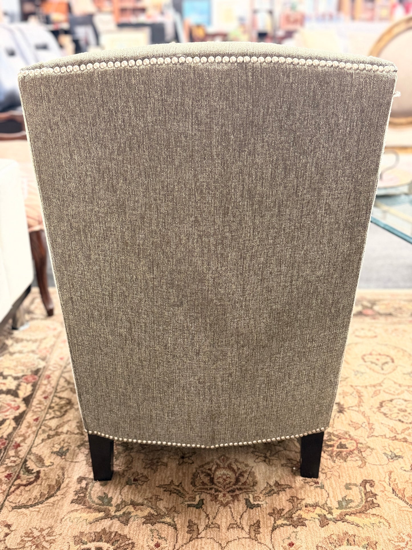 Gray upholstered armless accent chair featuring textured fabric, a gently curved tufted back, silver nailhead trim, and dark wooden legs.