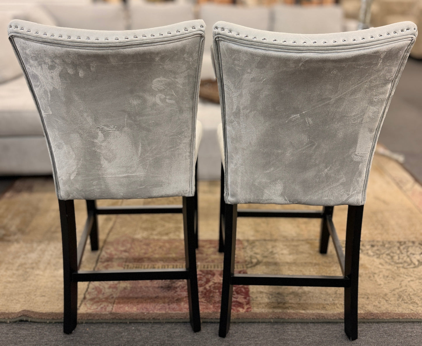 A pair of counter-height bar stools upholstered in light gray velvet-like fabric with silver nailhead trim and dark square wooden legs.