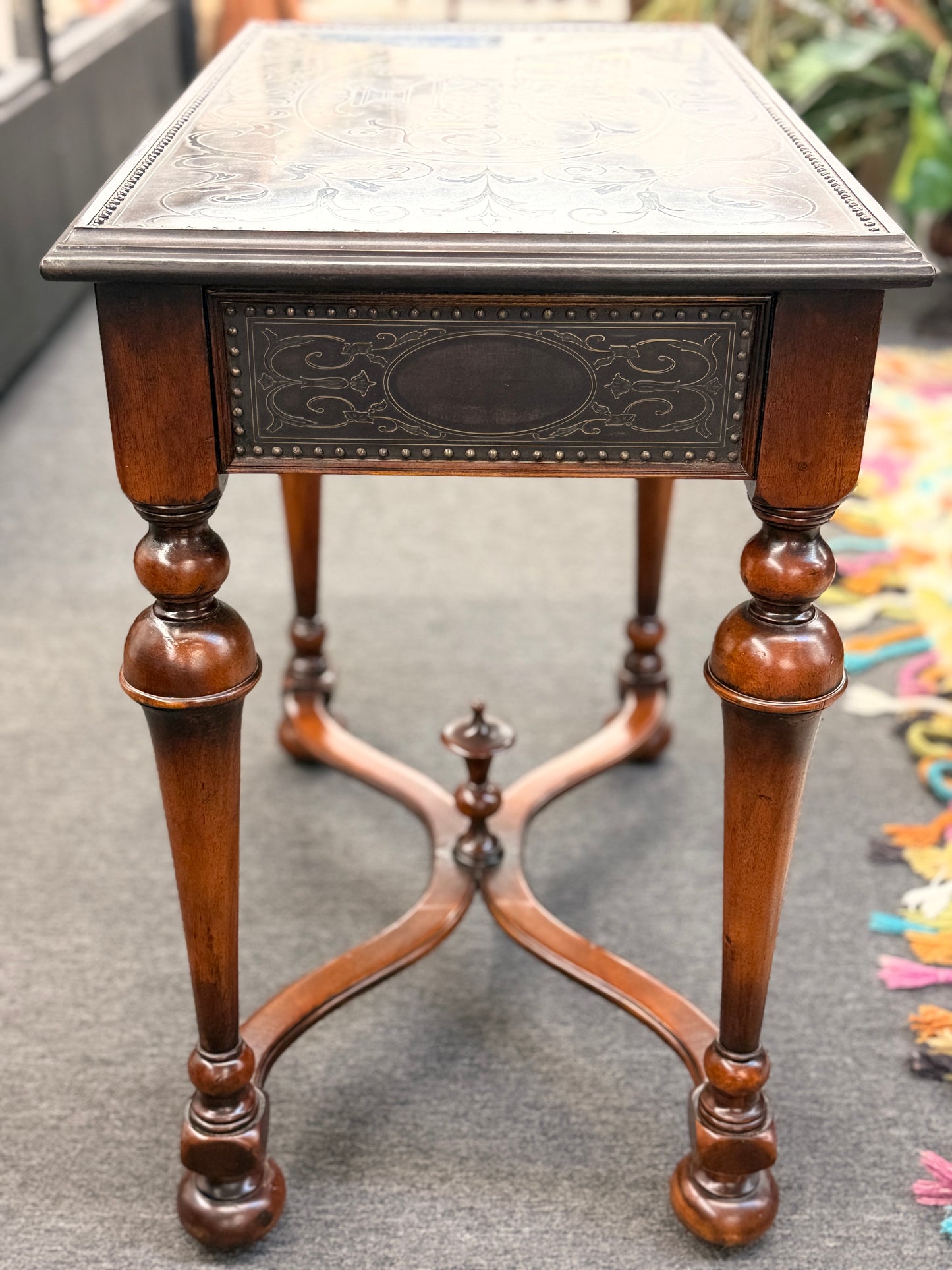 Traditional carved wood console table featuring two front drawers, turned carved legs, an embossed tabletop with nailhead trim, and a curved cross-stretcher with a decorative center finial.