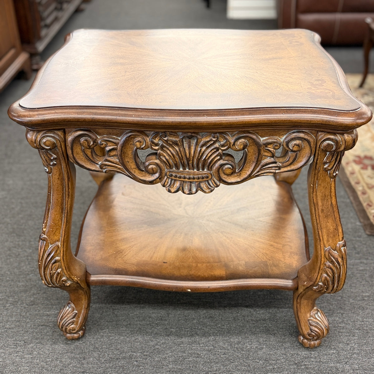 A traditional carved wood end table featuring ornate acanthus leaf aprons, cabriole legs, a sunburst wood veneer top, and a bottom display shelf in good condition.