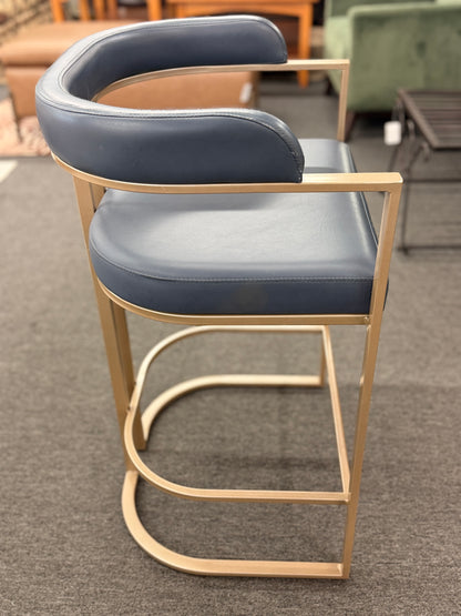 Modern bar stool with blue cushioned seat and gold frame on a gray floor.