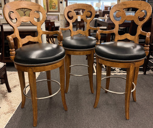 Three wooden bar stools with black leather seats in a showroom setting.