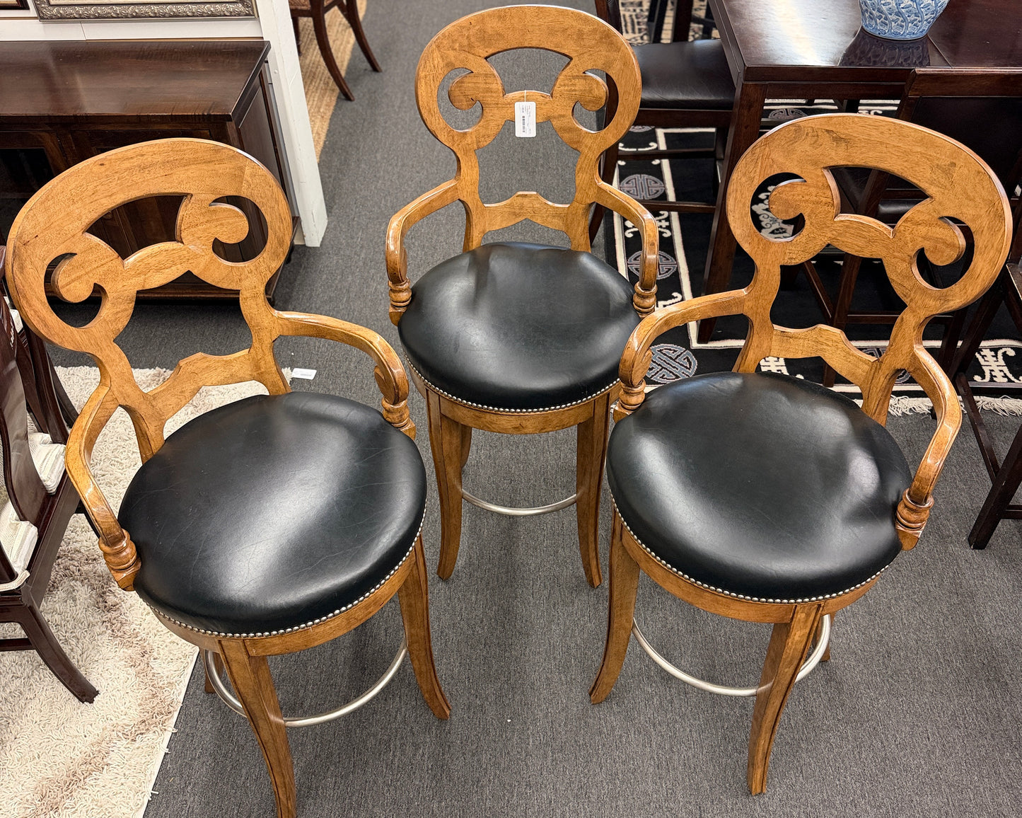 Three wooden chairs with black leather seats in a showroom setting.