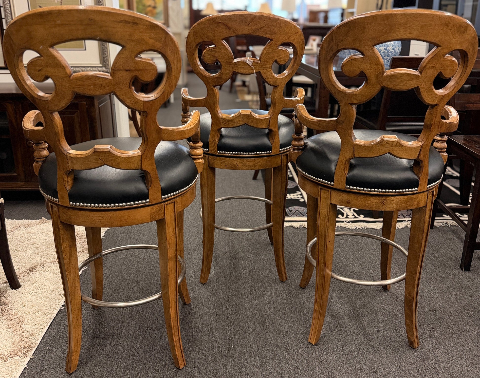 Three wooden bar stools with black leather seats in a showroom setting.