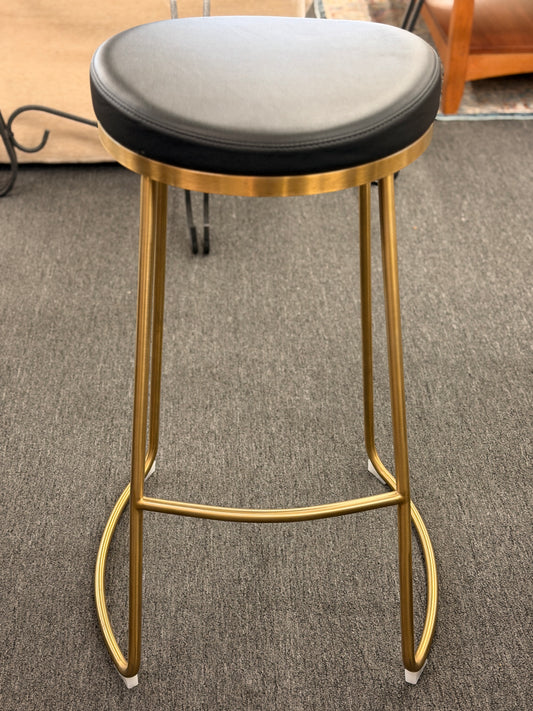 Bar stool with black cushioned seat and gold metal frame on a carpeted floor.