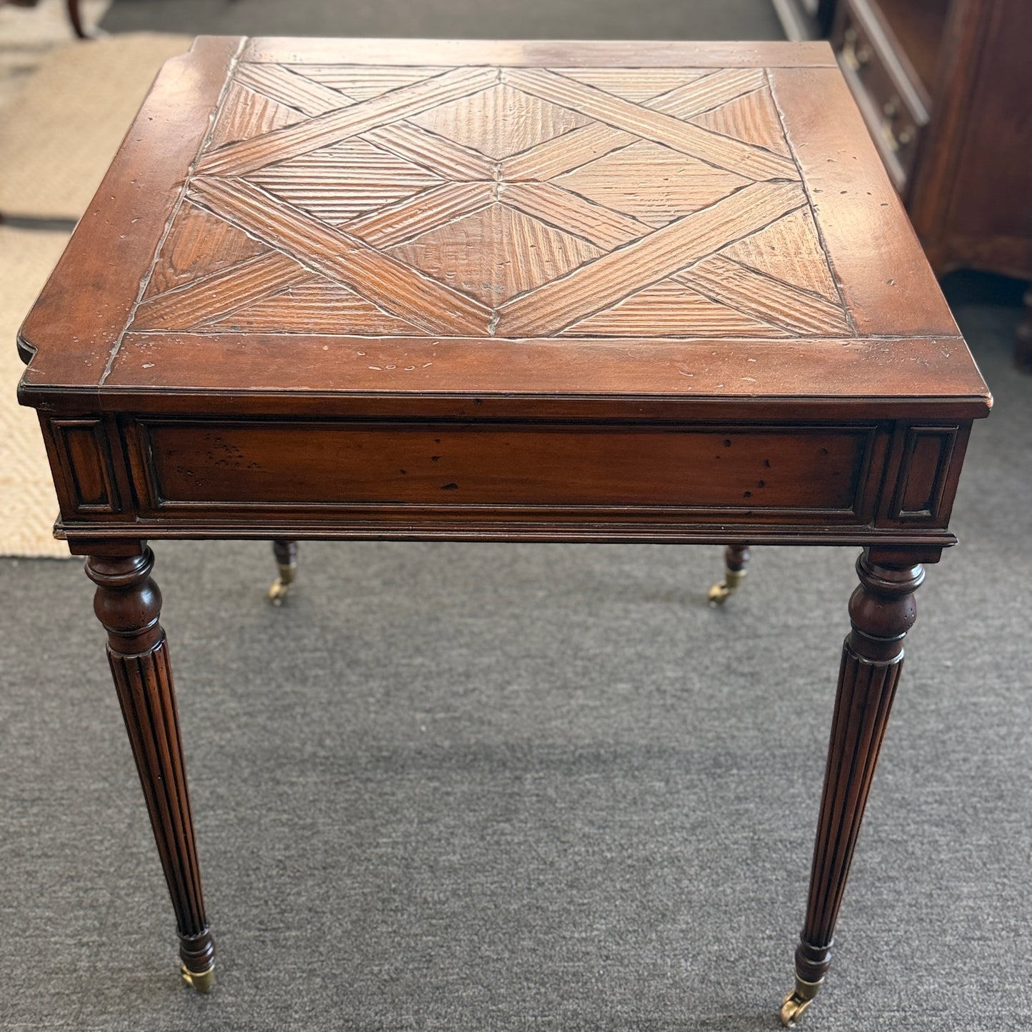A classic 26-inch square wood end table featuring a geometric parquet inlay top, one drawer with a brass ring pull, and fluted legs on small caster wheels.