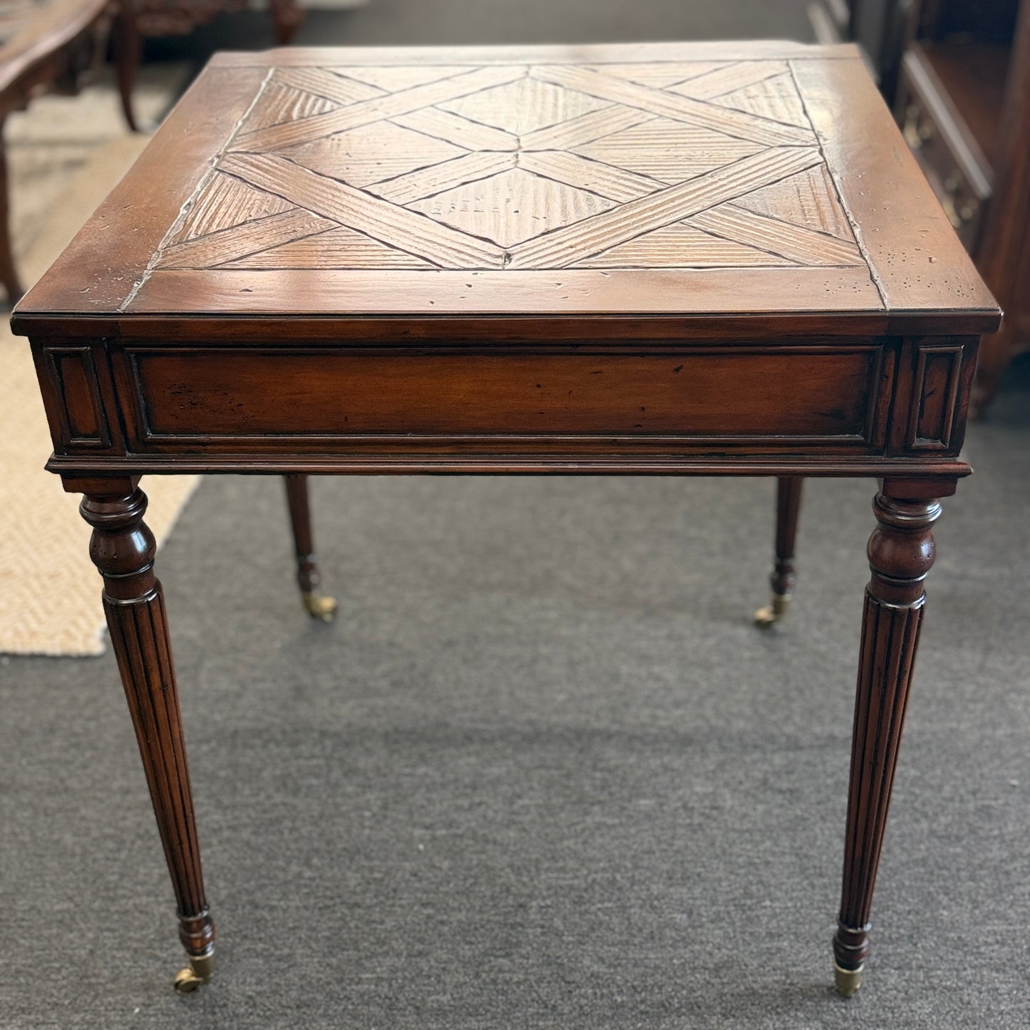 A classic 26-inch square wood end table featuring a geometric parquet inlay top, one drawer with a brass ring pull, and fluted legs on small caster wheels.