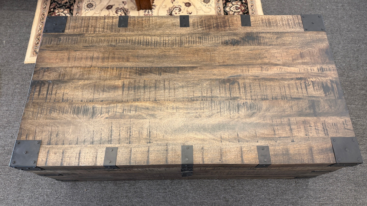 A rustic industrial trunk coffee table made of distressed dark wood with black iron corner brackets, open partially to reveal a lift-top mechanism and internal storage space.