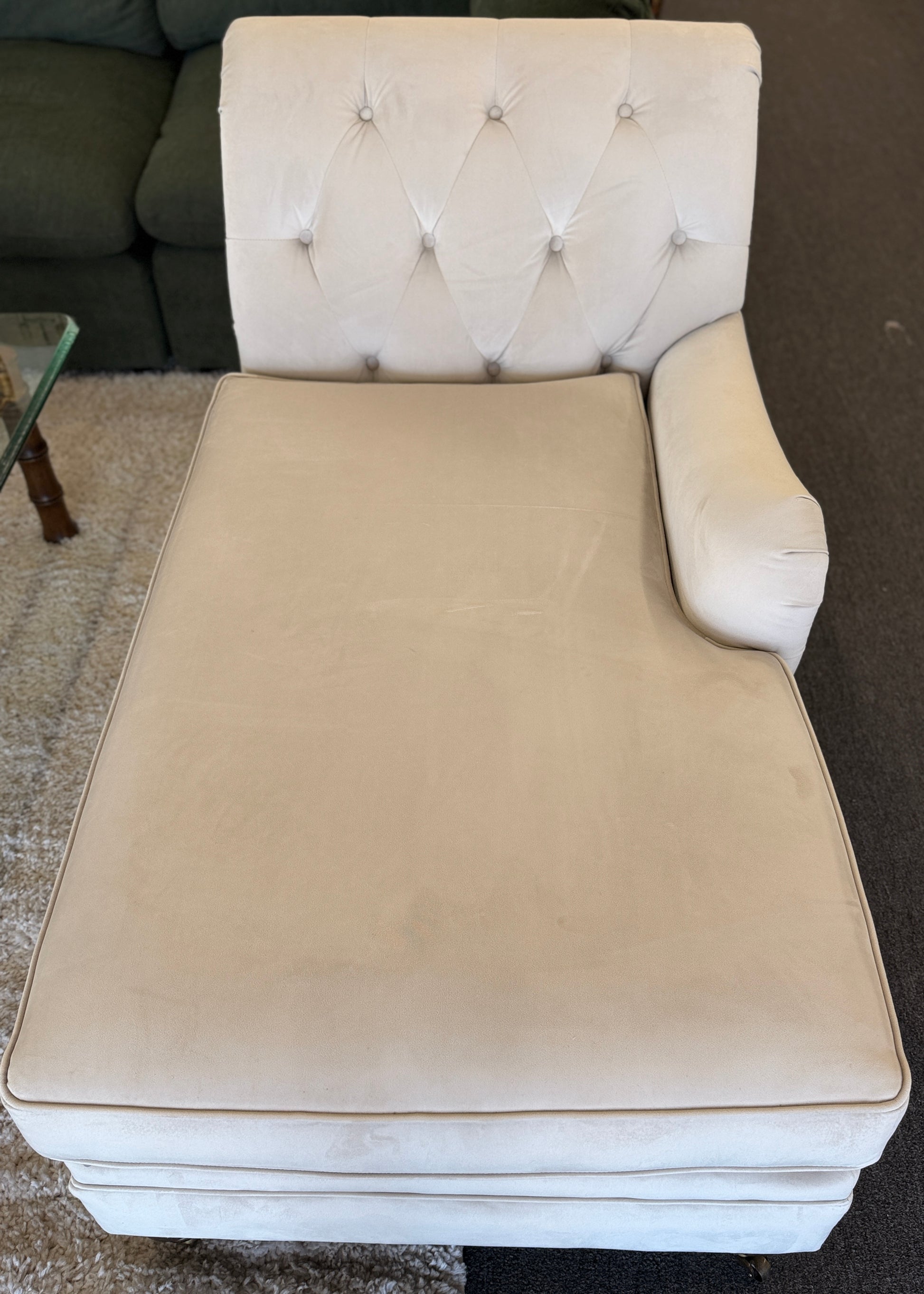 A vintage-style Bassett chaise lounge with a tufted rolled backrest, plush fabric cushion, and turned wooden legs on front casters.