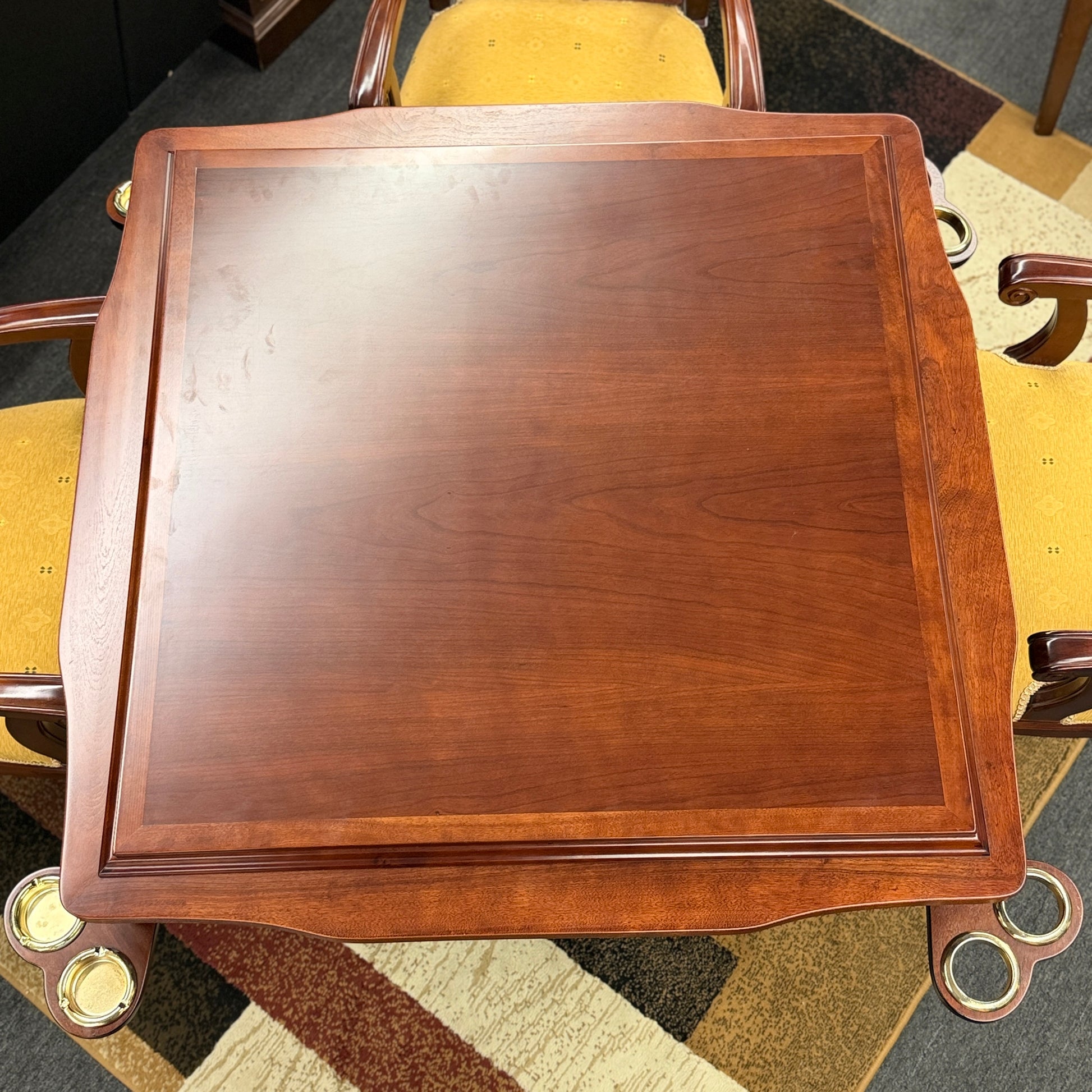 A 5-piece traditional game table set featuring a square wood table with a green felt surface and cup holders, surrounded by four rolling armchairs in yellow patterned upholstery with a Greek key trim.