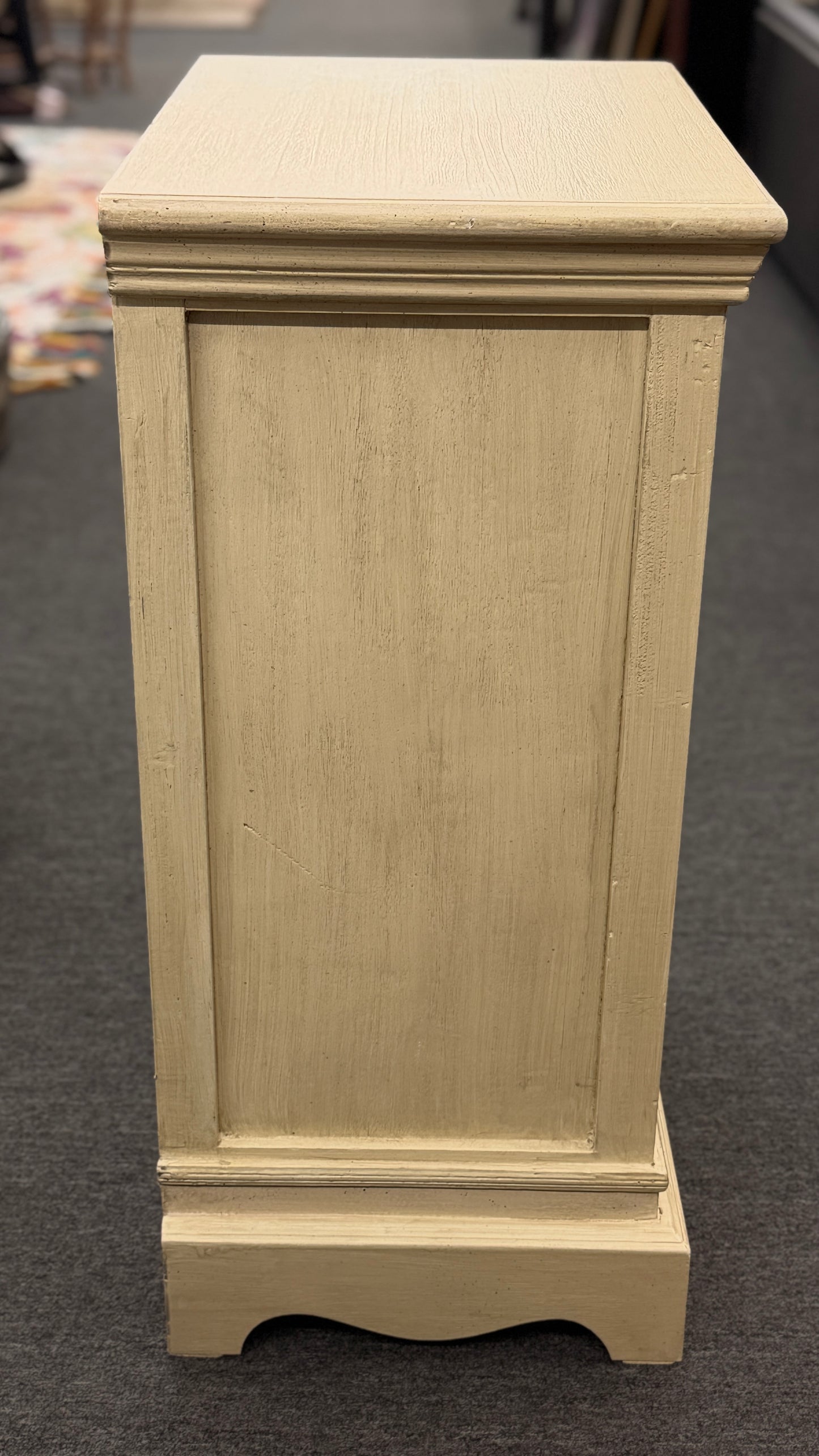 A small rustic accent cabinet with a distressed beige-cream painted finish, featuring a single louvered shutter door with a small dark round knob.