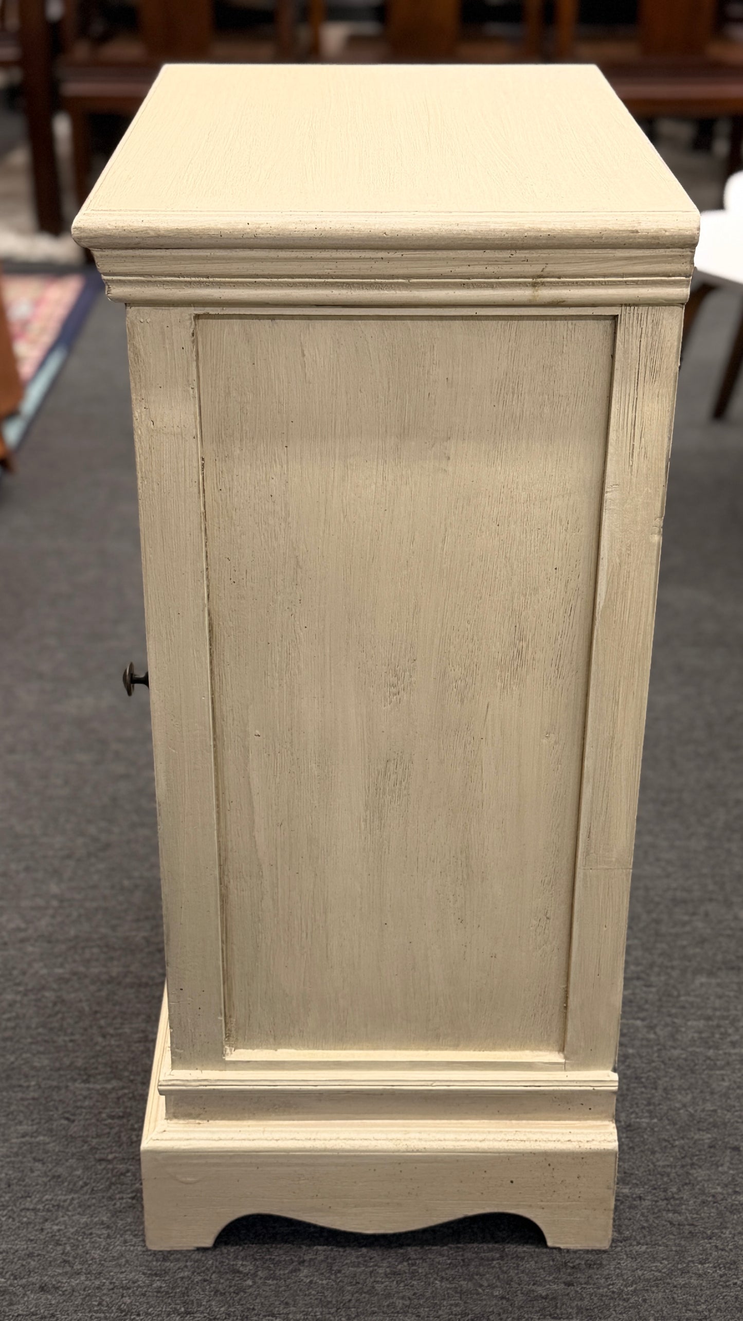 A small rustic accent cabinet with a distressed beige-cream painted finish, featuring a single louvered shutter door with a small dark round knob.