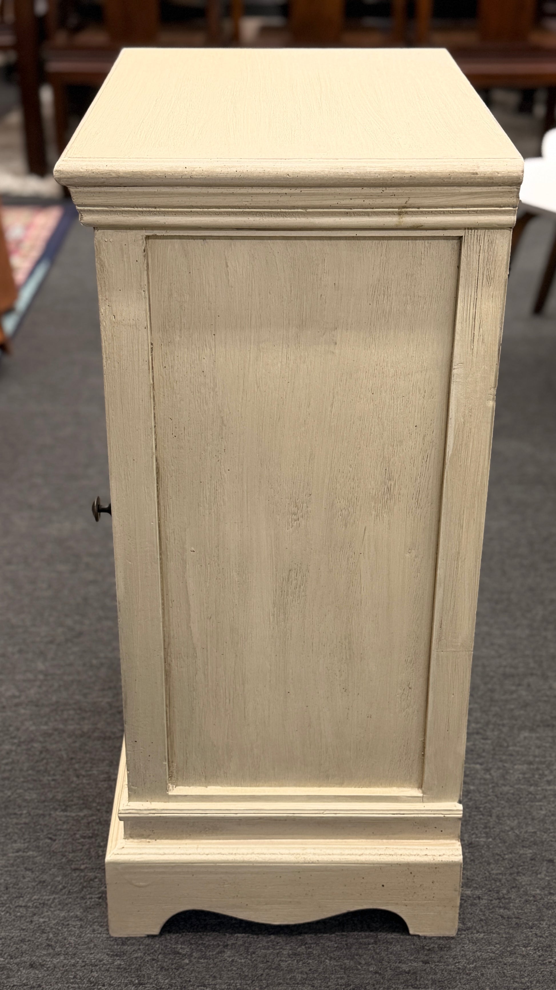 A small rustic accent cabinet with a distressed beige-cream painted finish, featuring a single louvered shutter door with a small dark round knob.