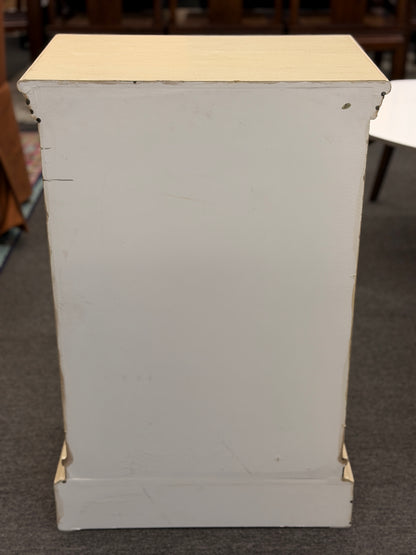 A small rustic accent cabinet with a distressed beige-cream painted finish, featuring a single louvered shutter door with a small dark round knob.