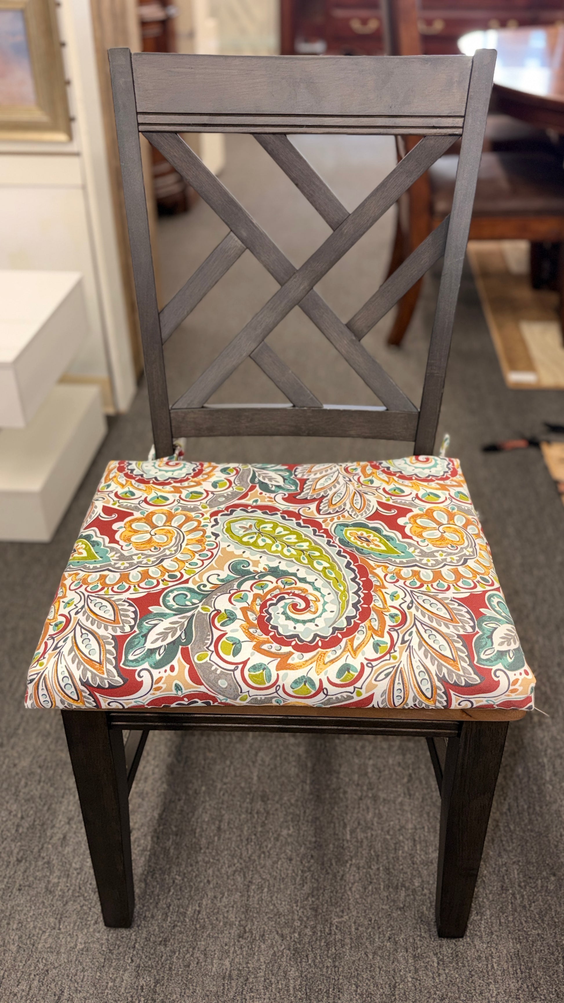 A casual oval dining table set featuring a mid-tone wood top, a dark base with an extendable leaf, and four matching X-back chairs with vibrant paisley-patterned tie-on cushions.
