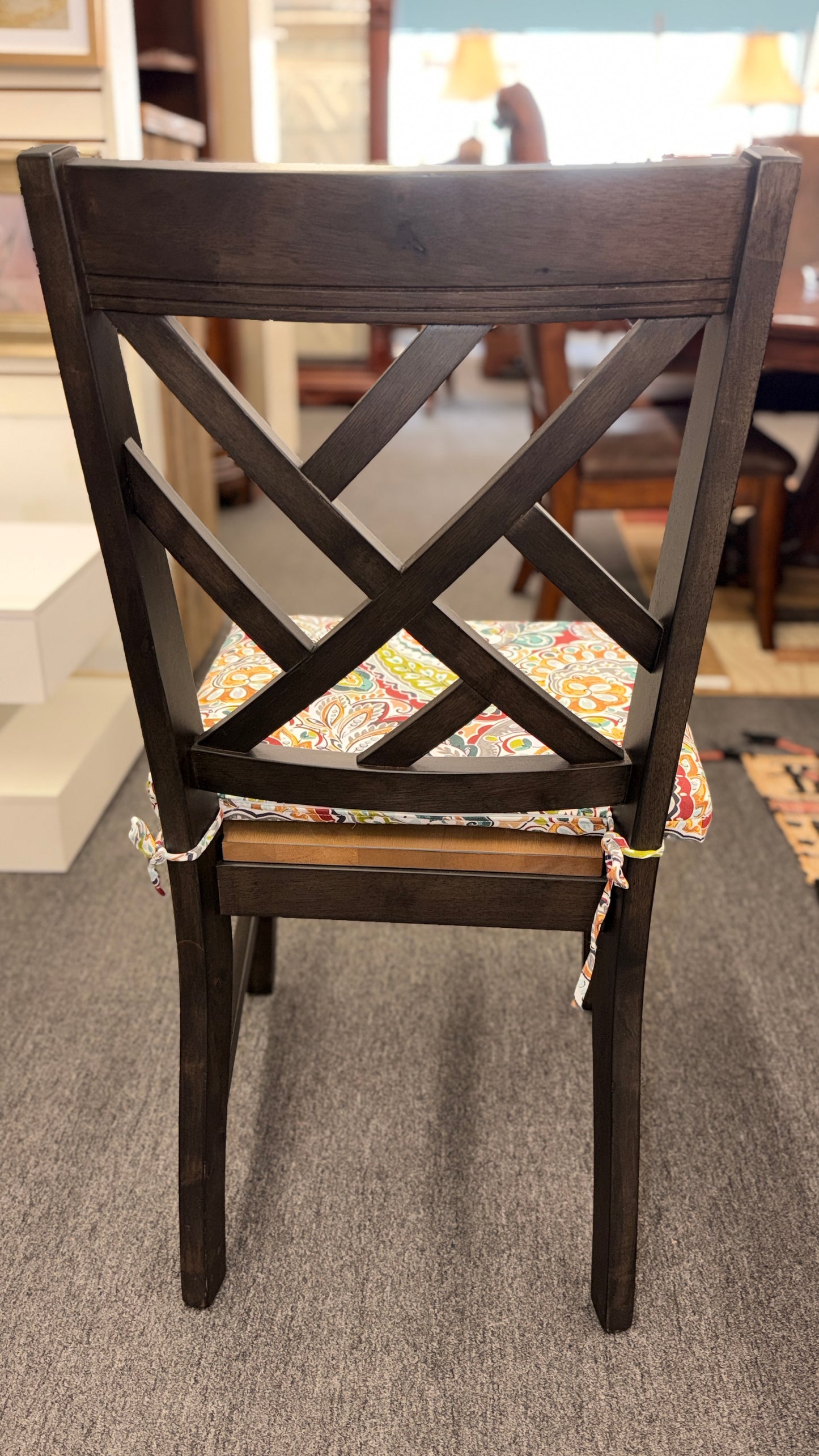 A casual oval dining table set featuring a mid-tone wood top, a dark base with an extendable leaf, and four matching X-back chairs with vibrant paisley-patterned tie-on cushions.