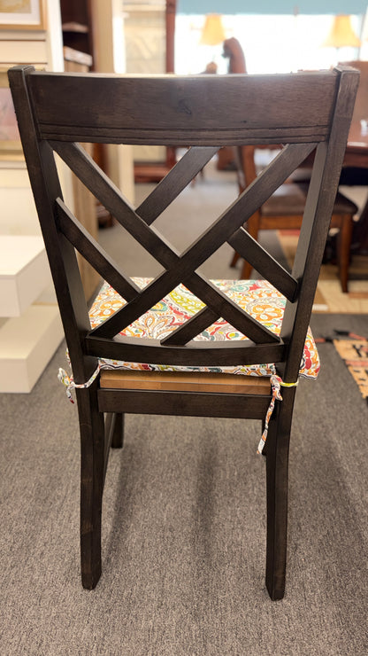 A casual oval dining table set featuring a mid-tone wood top, a dark base with an extendable leaf, and four matching X-back chairs with vibrant paisley-patterned tie-on cushions.