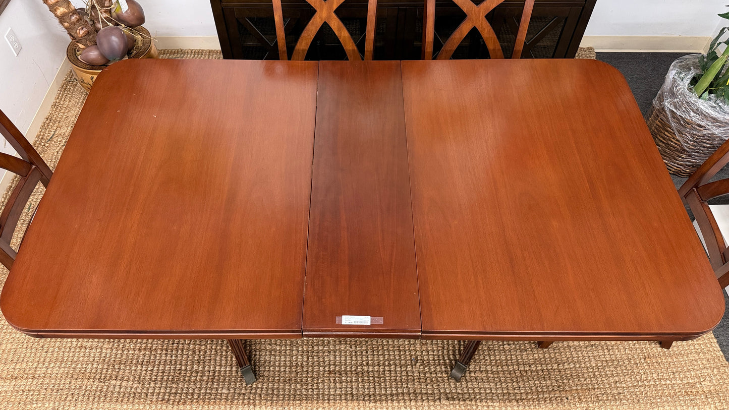 A traditional rectangular wooden dining table with an extendable leaf, twin turned pedestal bases, and six matching X-back dining chairs with light padded seats.