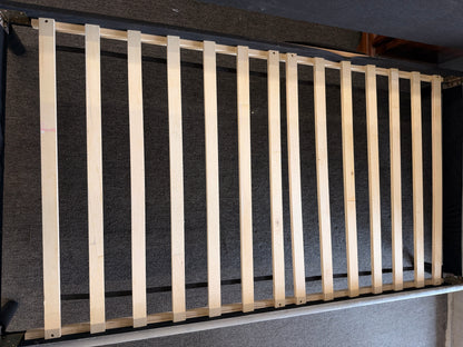A modern daybed frame upholstered in a gray textured fabric, featuring a mid-height backrest, enclosed side panels, and a wooden slatted mattress support base.