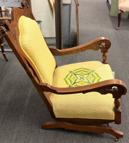 A vintage-style wooden platform rocking chair with an ornate carved crest rail, custom yellow upholstery, and a geometric green and blue embroidered center design.