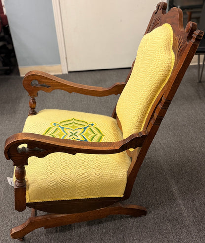 A vintage-style wooden platform rocking chair with an ornate carved crest rail, custom yellow upholstery, and a geometric green and blue embroidered center design.