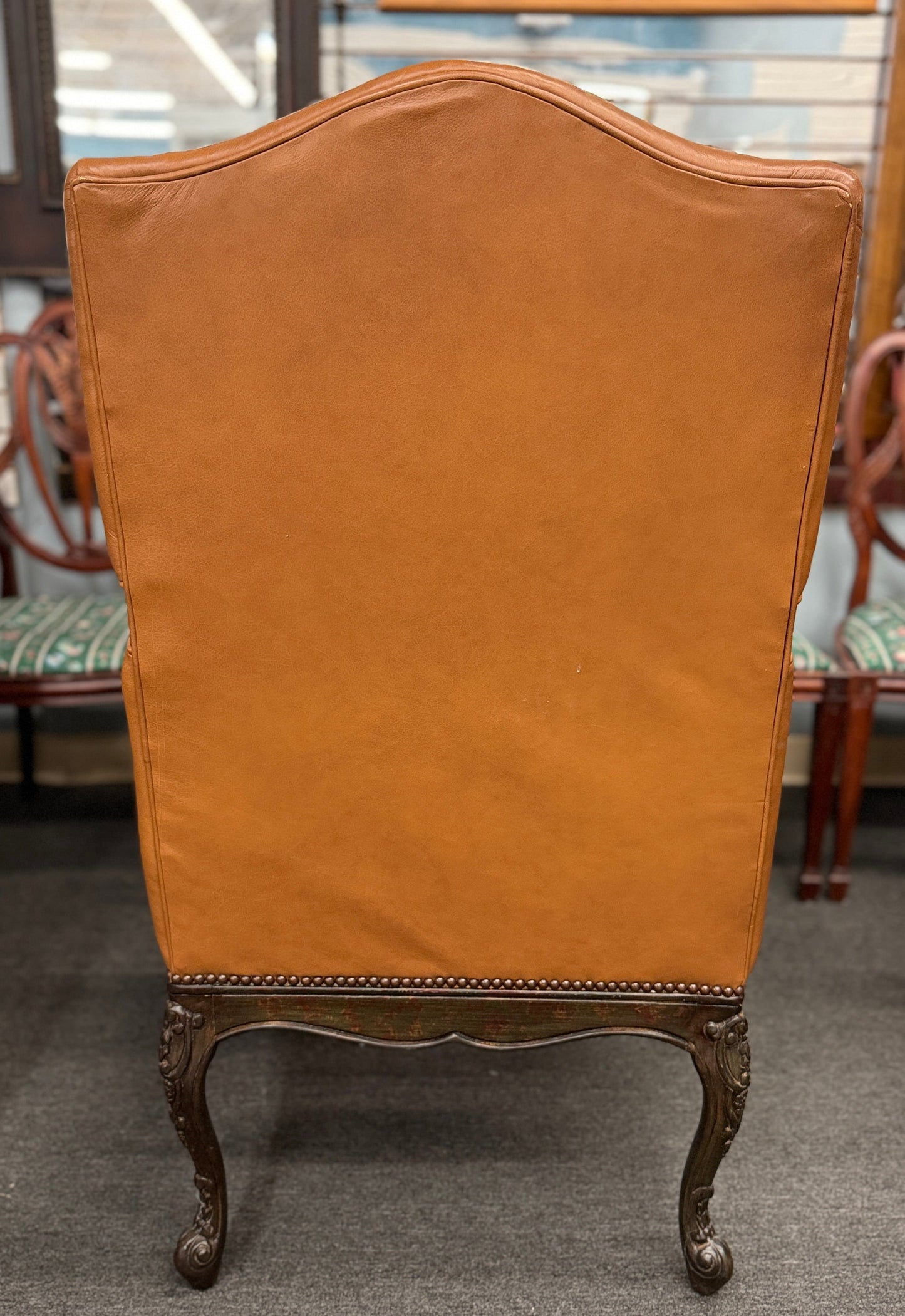 An elegant antique-reproduction accent chair upholstered in smooth tan leather with a hand-carved dark wood frame, cabriole legs, and brass nailhead trim.