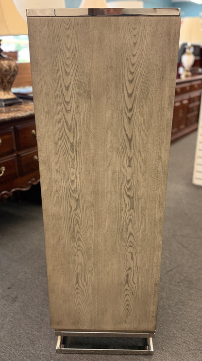 A contemporary tall 7-drawer highboy chest of drawers with a soft gray-washed wood grain finish, minimalist silver hardware, and a geometric chrome base.