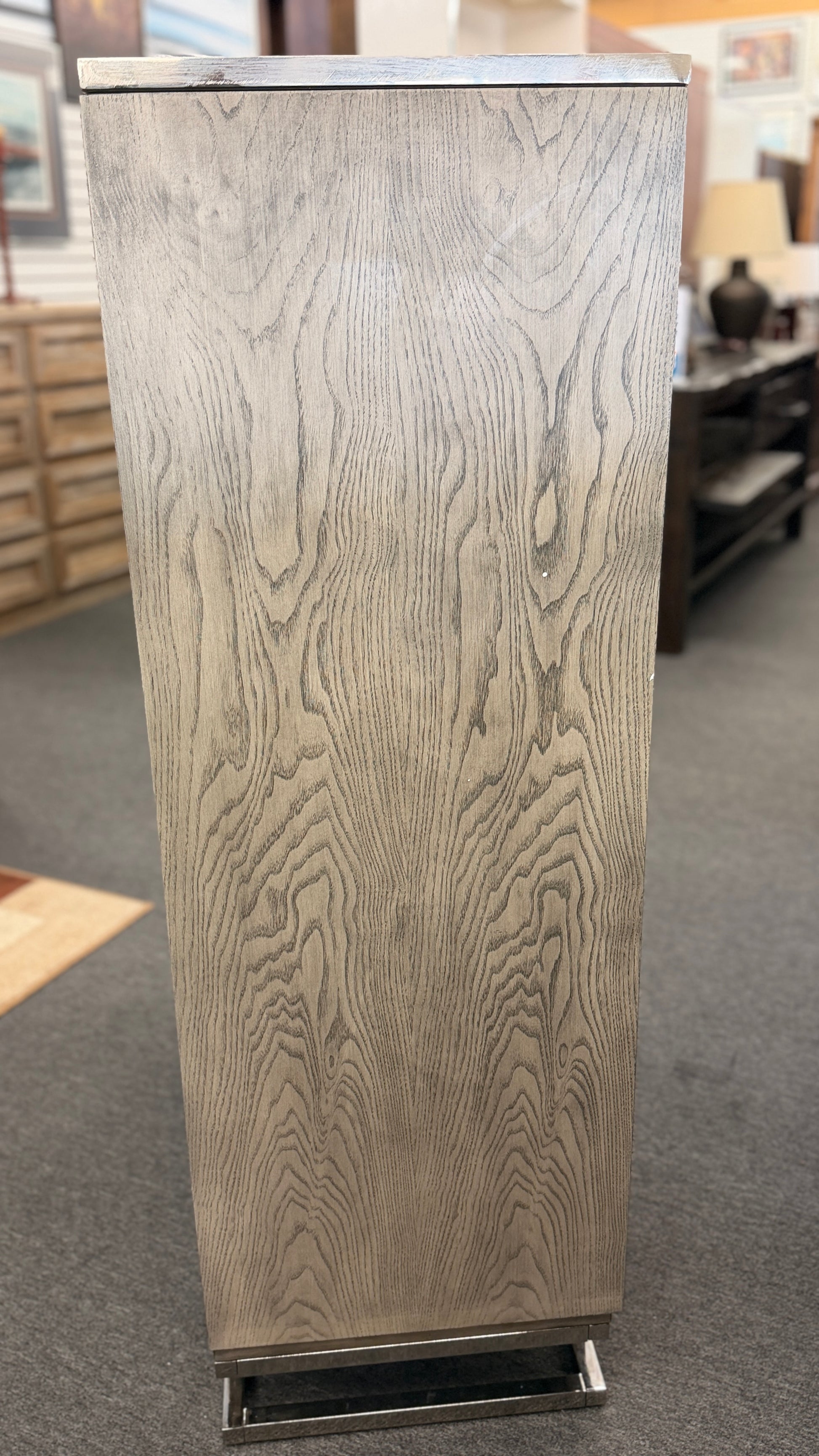 A contemporary tall 7-drawer highboy chest of drawers with a soft gray-washed wood grain finish, minimalist silver hardware, and a geometric chrome base.