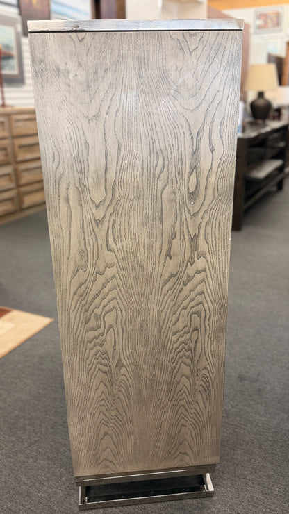 A contemporary tall 7-drawer highboy chest of drawers with a soft gray-washed wood grain finish, minimalist silver hardware, and a geometric chrome base.