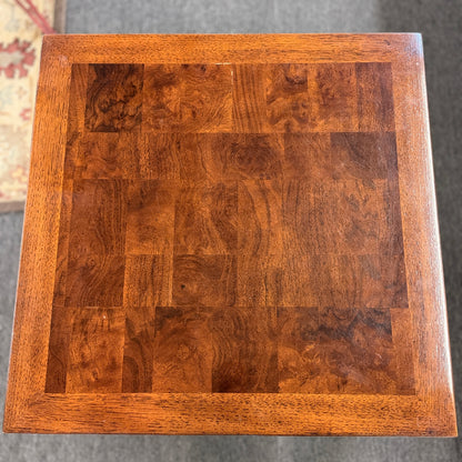 A modern square accent table with thick blocky legs flush with the corners and a highly reflective, glossy top surface showing a rich burl wood pattern.