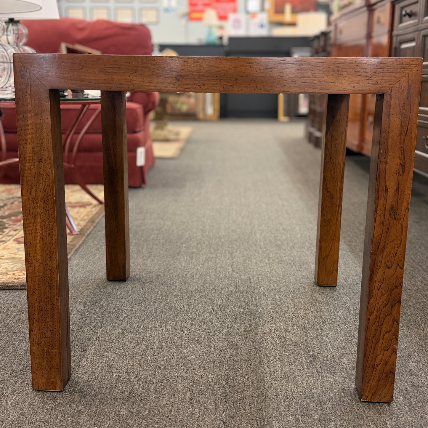 A modern square accent table with thick blocky legs flush with the corners and a highly reflective, glossy top surface showing a rich burl wood pattern.