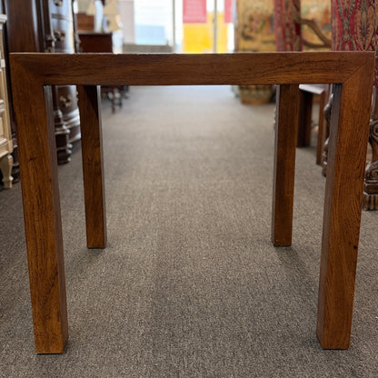 A modern square accent table with thick blocky legs flush with the corners and a highly reflective, glossy top surface showing a rich burl wood pattern.