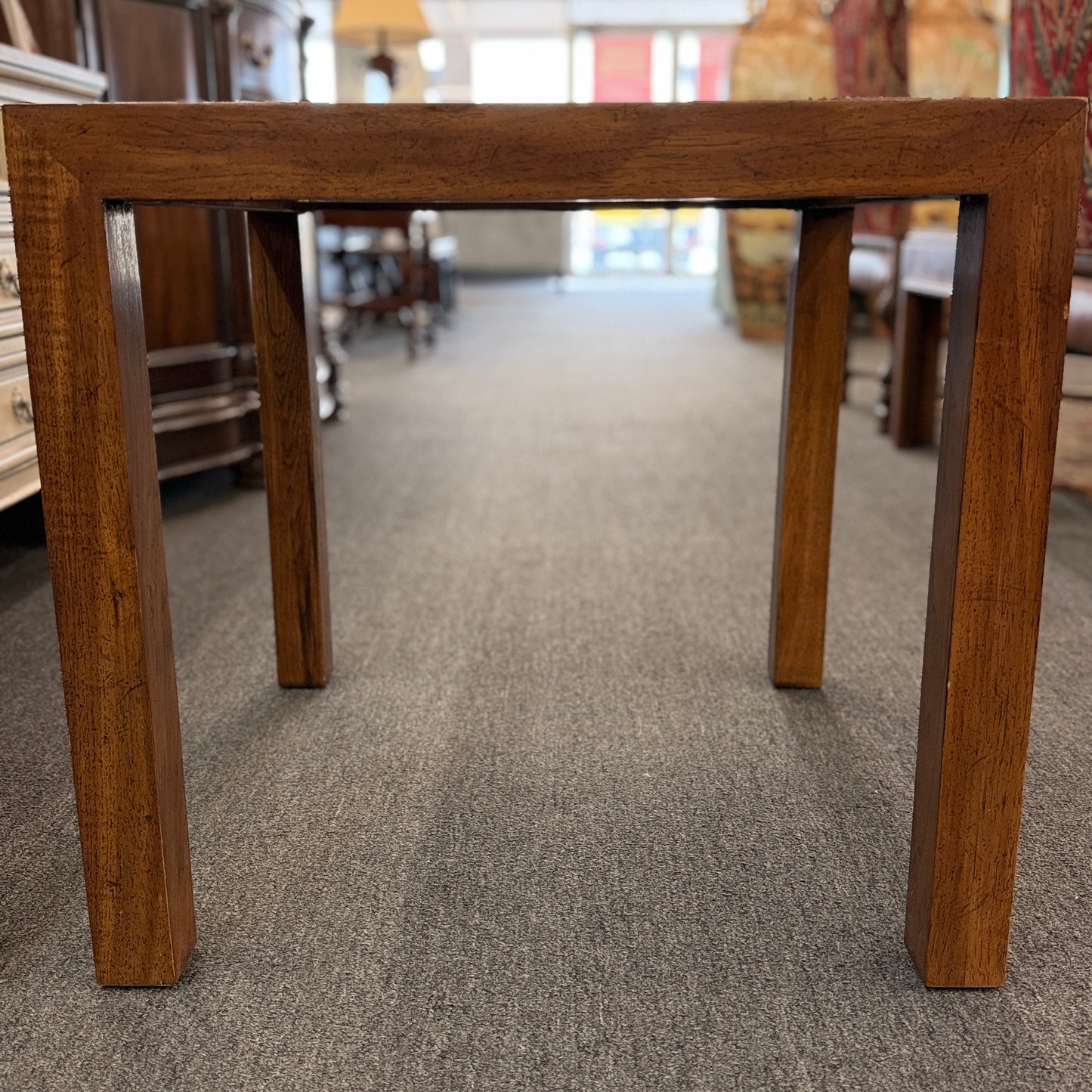 A modern square accent table with thick blocky legs flush with the corners and a highly reflective, glossy top surface showing a rich burl wood pattern.