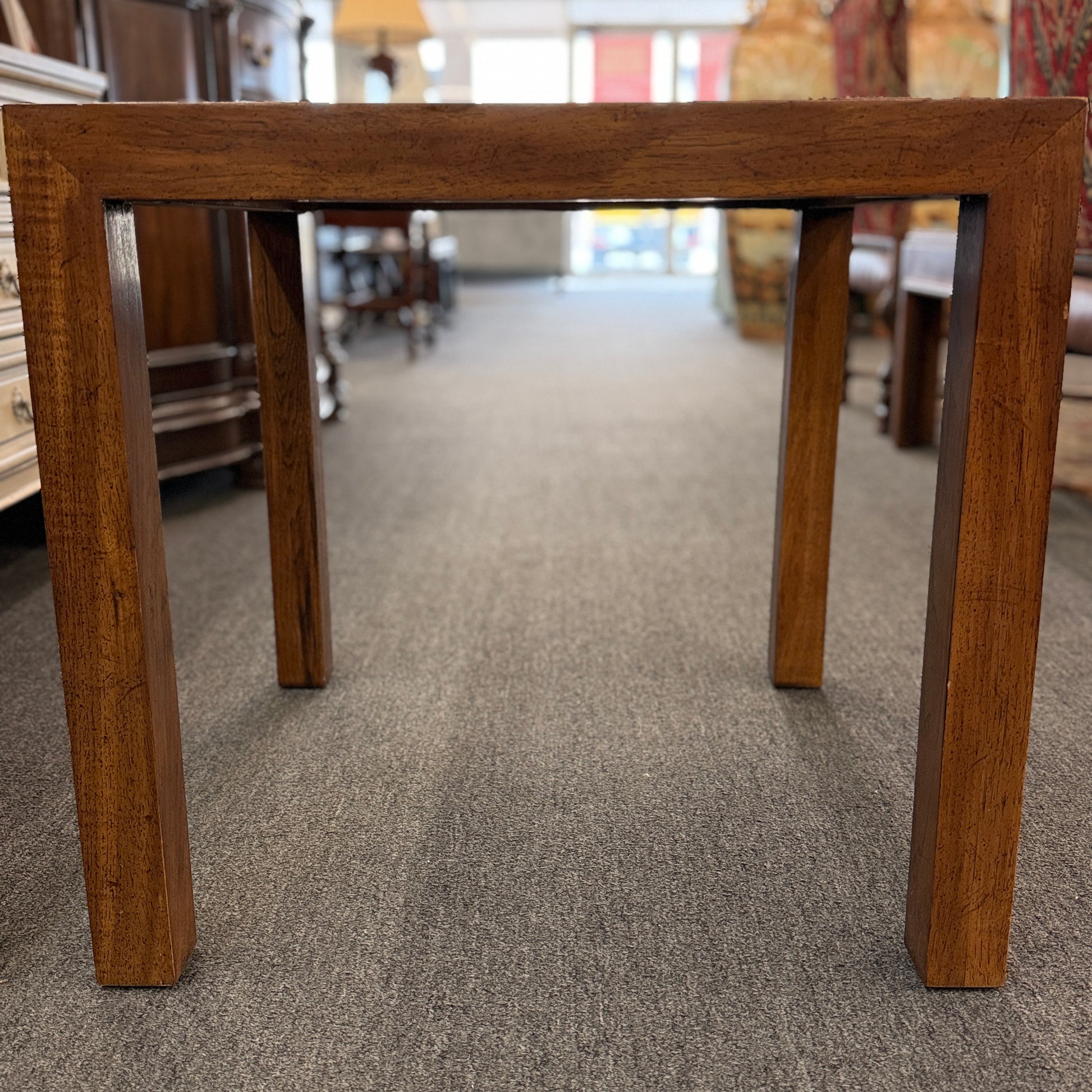 A modern square accent table with thick blocky legs flush with the corners and a highly reflective, glossy top surface showing a rich burl wood pattern.