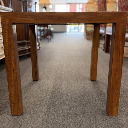 A modern square accent table with thick blocky legs flush with the corners and a highly reflective, glossy top surface showing a rich burl wood pattern.
