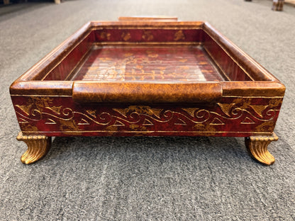 A rectangular wooden decorative vanity tray with ornate gold wave patterns on the sides, handles, short feet, and a faux-tortoiseshell interior with gold botanical designs.