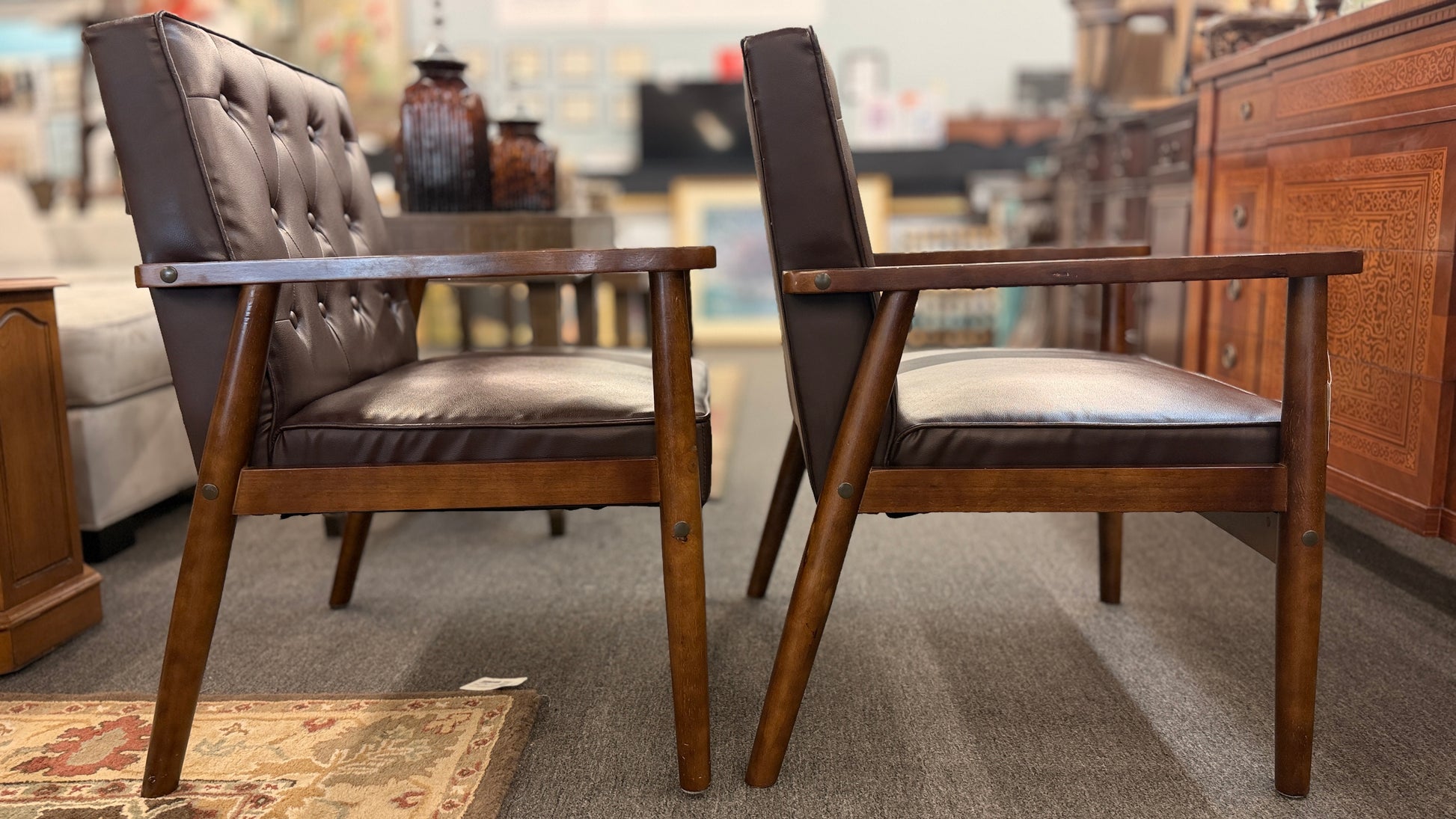 A pair of mid-century modern style accent armchairs featuring dark brown button-tufted backrests, smooth seat cushions, and exposed medium-toned wooden frames with tapered legs.