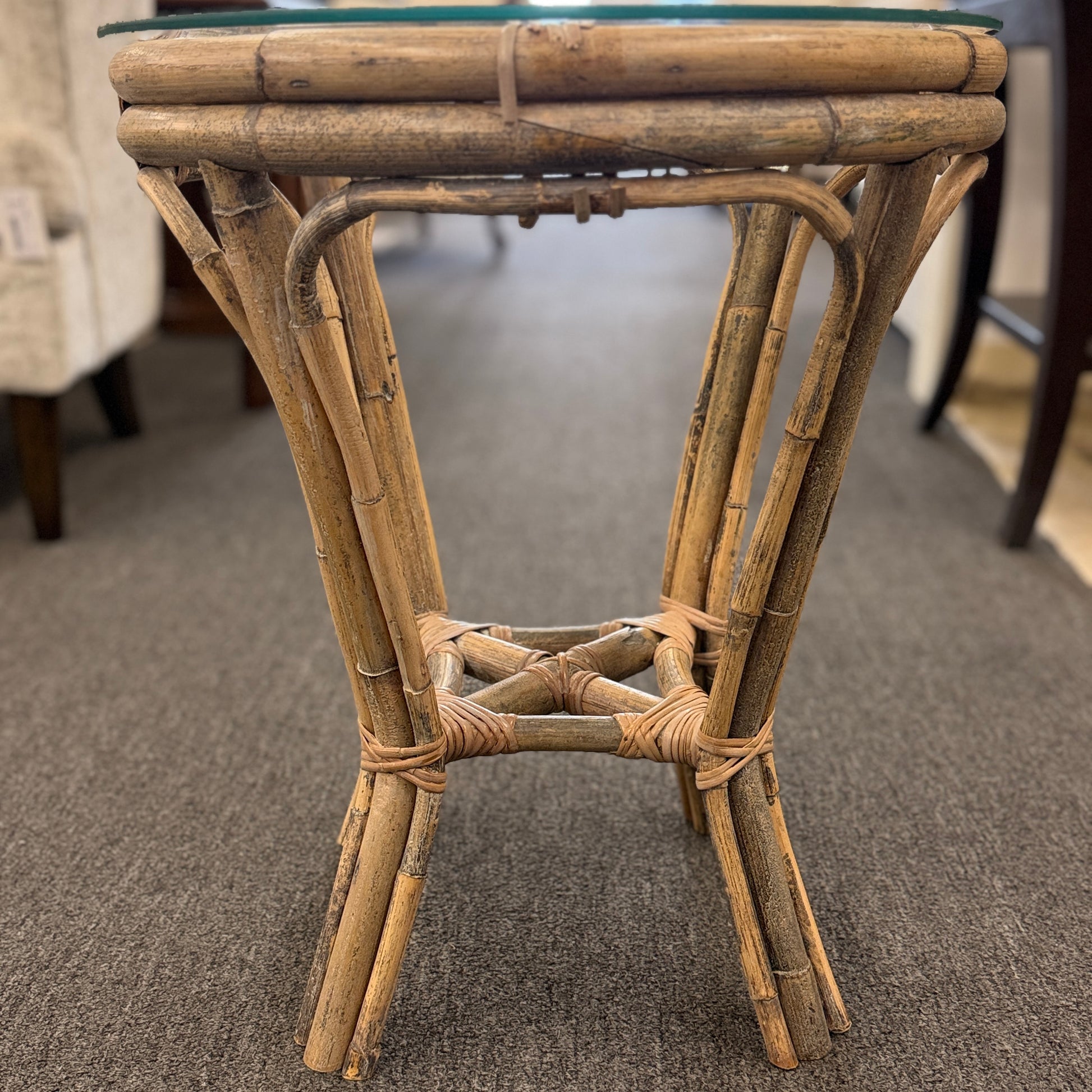 A small round end table with a bent organic-style bamboo frame, wrapped structural joints, an X-shaped cross-stretcher, and a clear glass tabletop.