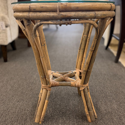 A small round end table with a bent organic-style bamboo frame, wrapped structural joints, an X-shaped cross-stretcher, and a clear glass tabletop.