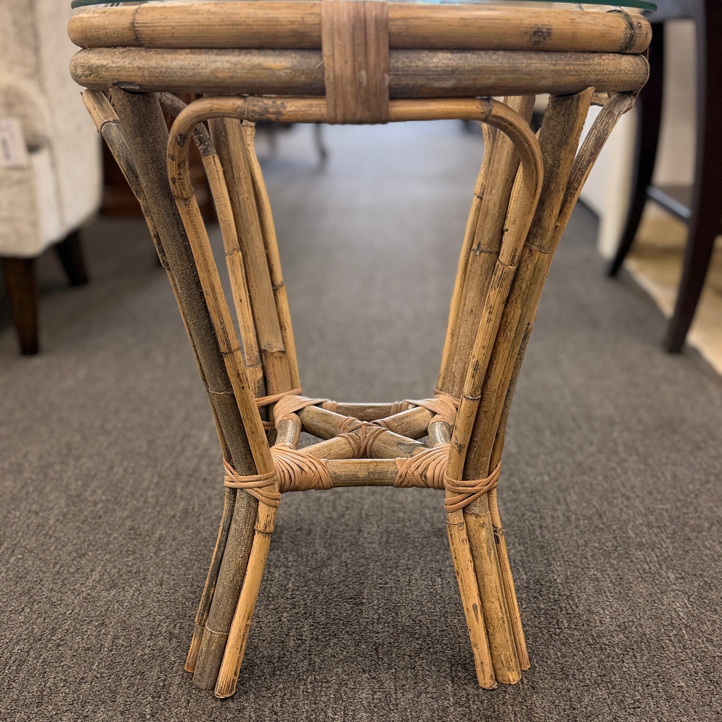A small round end table with a bent organic-style bamboo frame, wrapped structural joints, an X-shaped cross-stretcher, and a clear glass tabletop.
