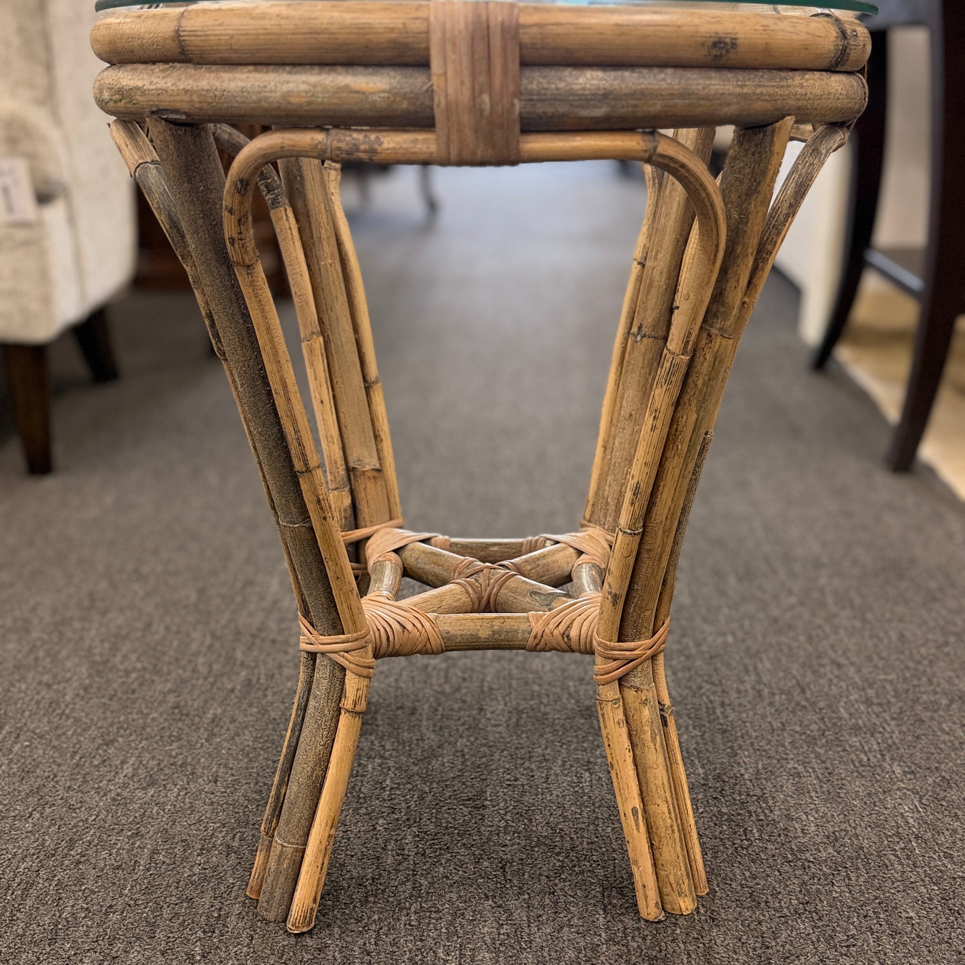 A small round end table with a bent organic-style bamboo frame, wrapped structural joints, an X-shaped cross-stretcher, and a clear glass tabletop.