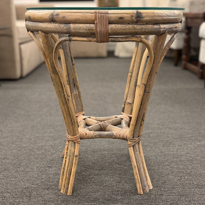 A small round end table with a bent organic-style bamboo frame, wrapped structural joints, an X-shaped cross-stretcher, and a clear glass tabletop.
