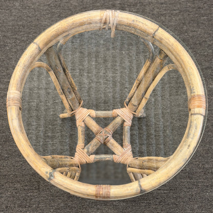 A small round end table with a bent organic-style bamboo frame, wrapped structural joints, an X-shaped cross-stretcher, and a clear glass tabletop.