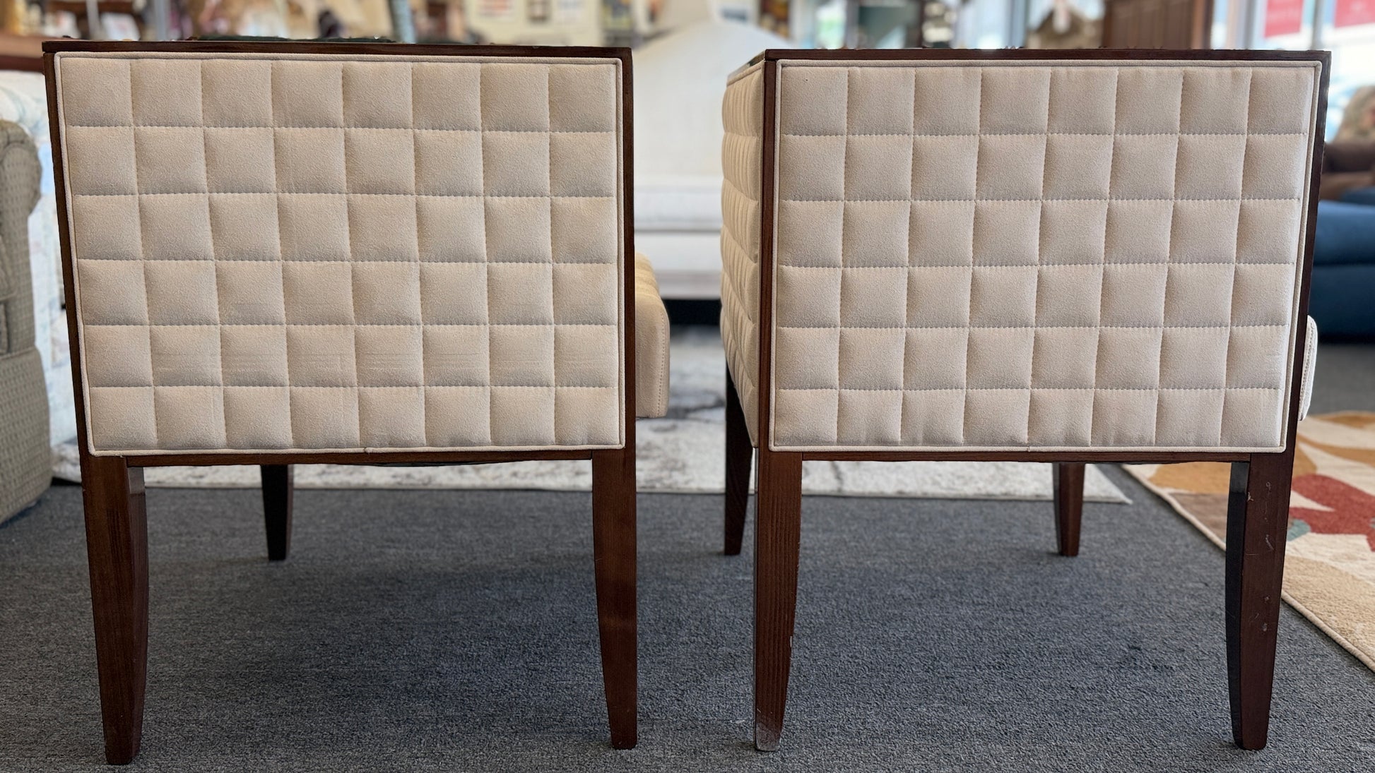 A pair of contemporary squared-off accent chairs with light beige suede-like upholstery, an exposed dark wood outer frame, and subtle quilted tufting on the side panels.