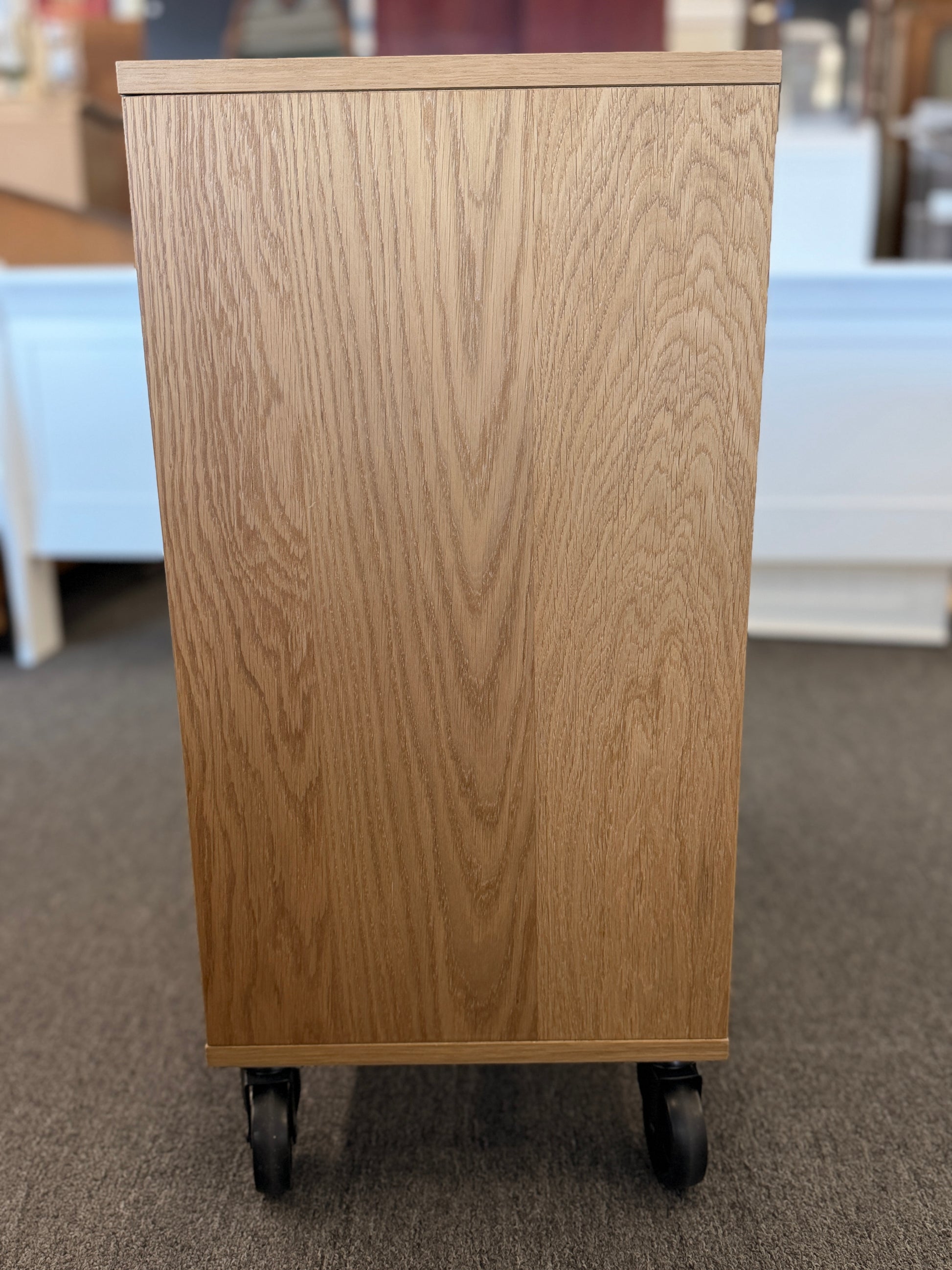 A modern mobile office storage cart with a light oak finish, featuring asymmetrical divided cubby compartments and four black rolling casters.